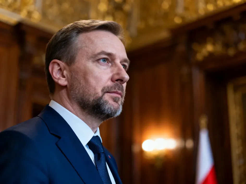 Elegancki mężczyzna w garniturze w historycznym wnętrzu z polską flagą w tle, nawiązujący do powagi urzędu prezydenta.
